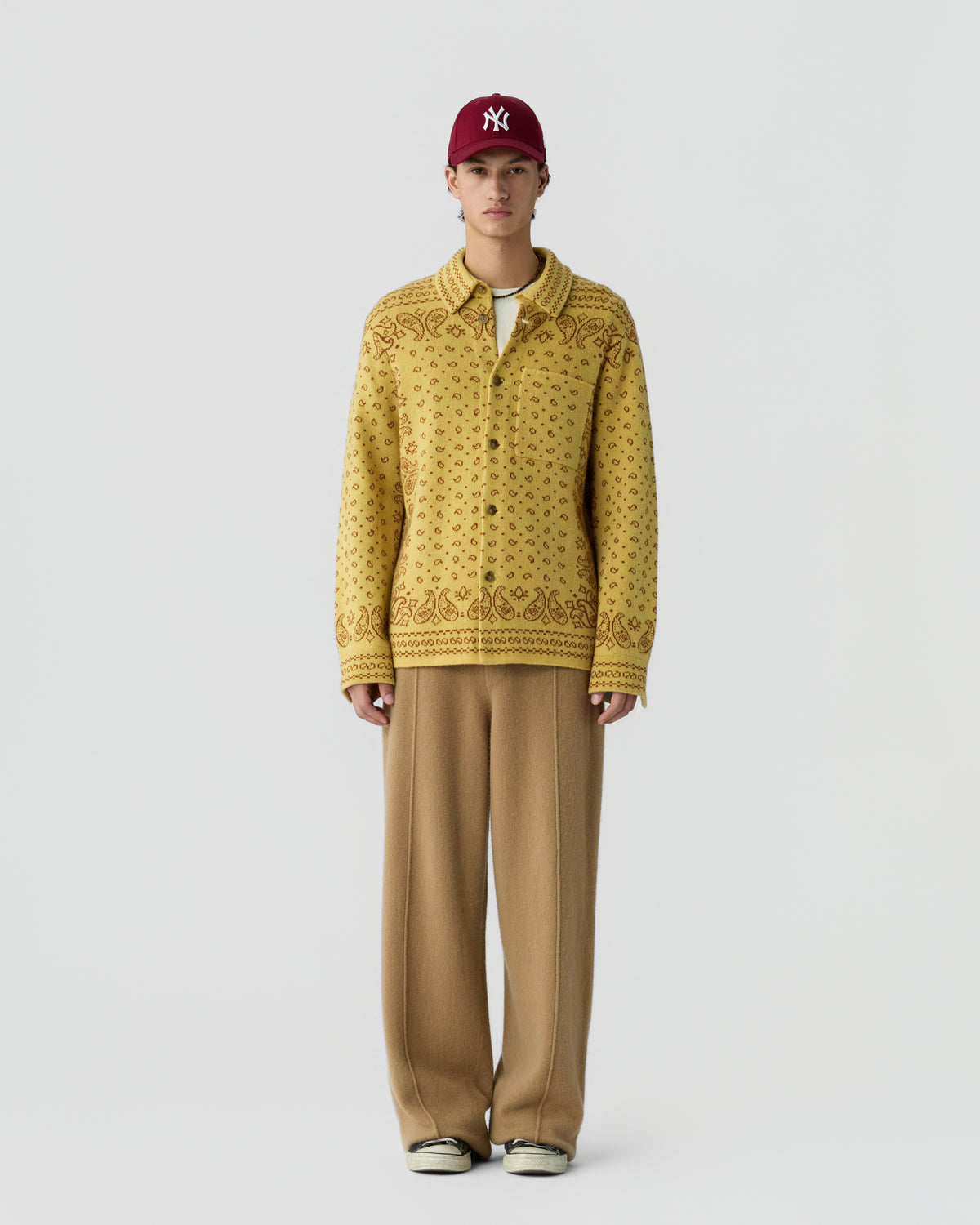 Young man wearing a patterned yellow button-up cardigan, wide-legged brown trousers, a maroon cap, and white sneakers, standing against a plain white background.