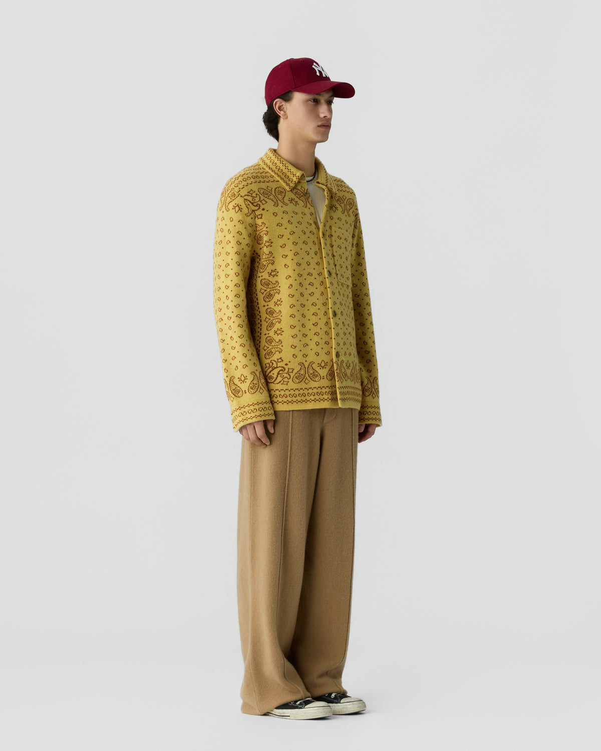 A young man wearing a red baseball cap, a yellow patterned zip-up sweater, wide beige pants, and black and white sneakers, standing against a plain white background.