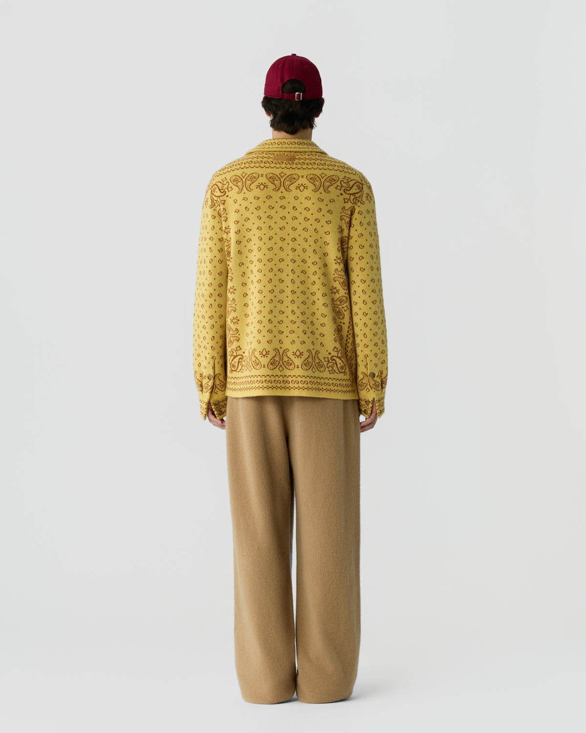 Person wearing a yellow patterned long-sleeve sweater, wide beige pants, and a red cap, standing facing away on a plain light gray background.