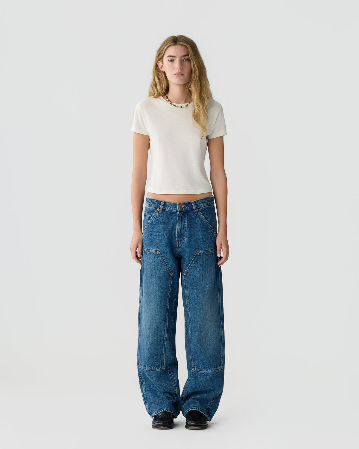 Full-length view of a woman wearing a cream short-sleeved top, relaxed blue jeans, and black shoes against a light gray background