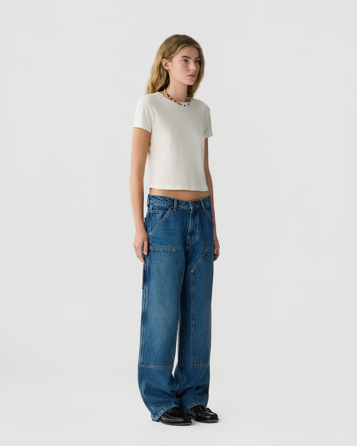 Woman wearing a white fitted T shirt, loose blue jeans, and black shoes while standing against a light gray studio background.