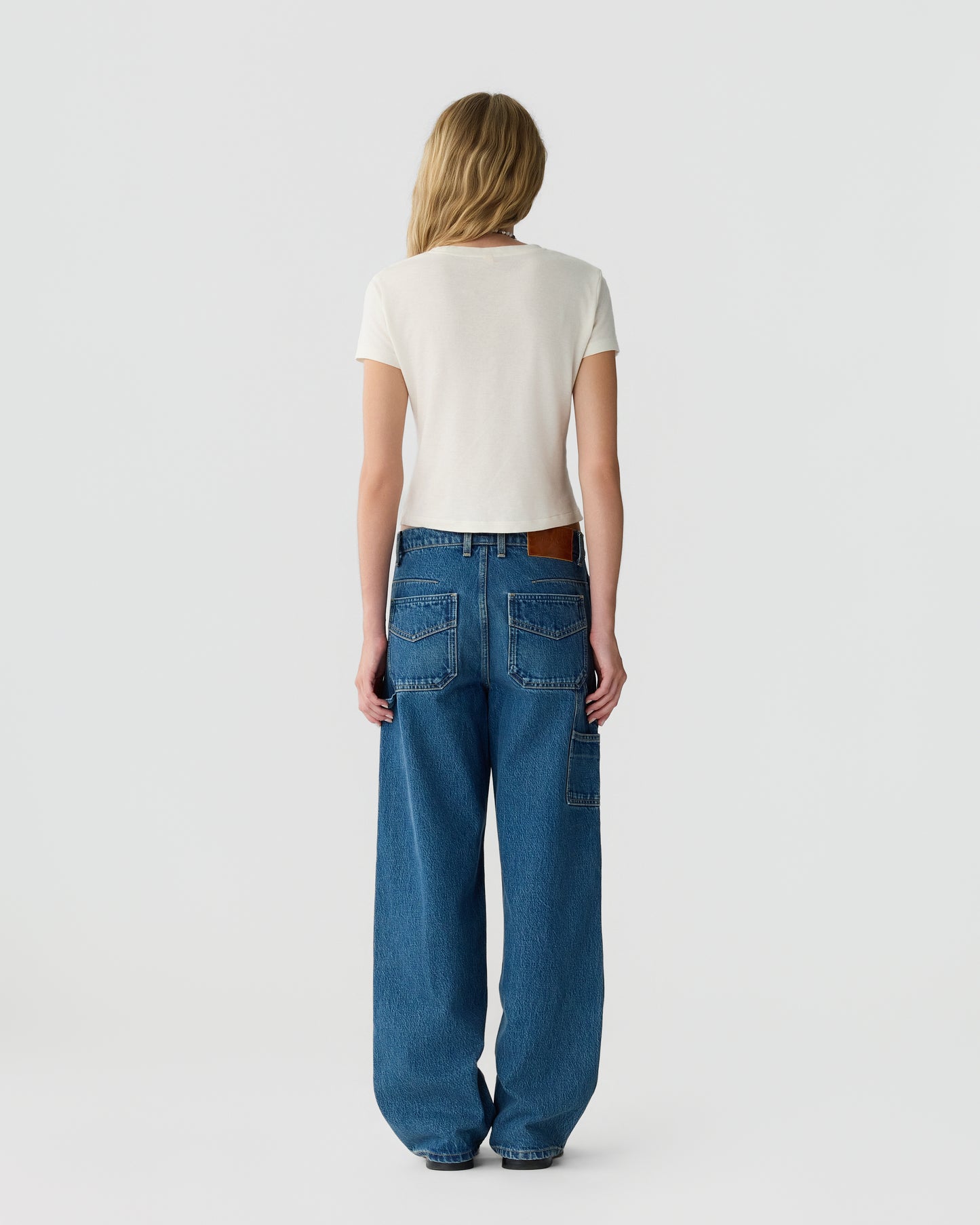 Back view of a woman wearing a white short sleeve top and relaxed blue jeans with multiple pockets against a plain light background